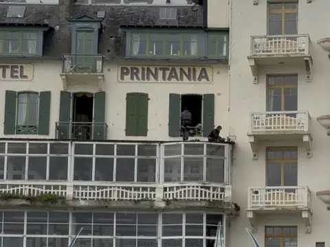 Deux couvreurs intervenant sur la corniche et le bow-window d'un hôtel du front de mer, façade à volets verts