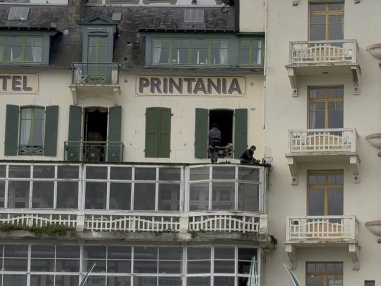 Deux couvreurs intervenant sur la corniche et le bow-window d'un hôtel du front de mer, façade à volets verts