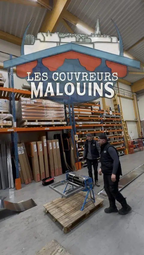 À l'atelier de Saint-Malo : deux couvreurs devant la plieuse posée sur une palette, feuilles de zinc et rayonnages en arrière-plan