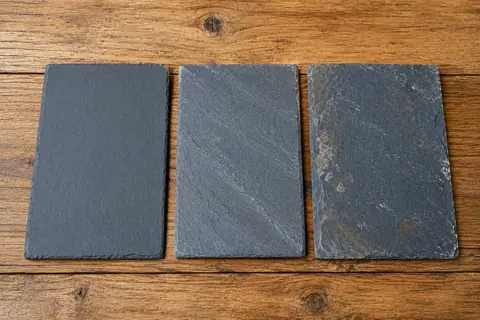 Trois échantillons d'ardoise naturelle côte à côte sur une table en bois : premier choix lisse et régulier, deuxième choix à la texture plus marquée, troisième choix avec inclusions et bords irréguliers