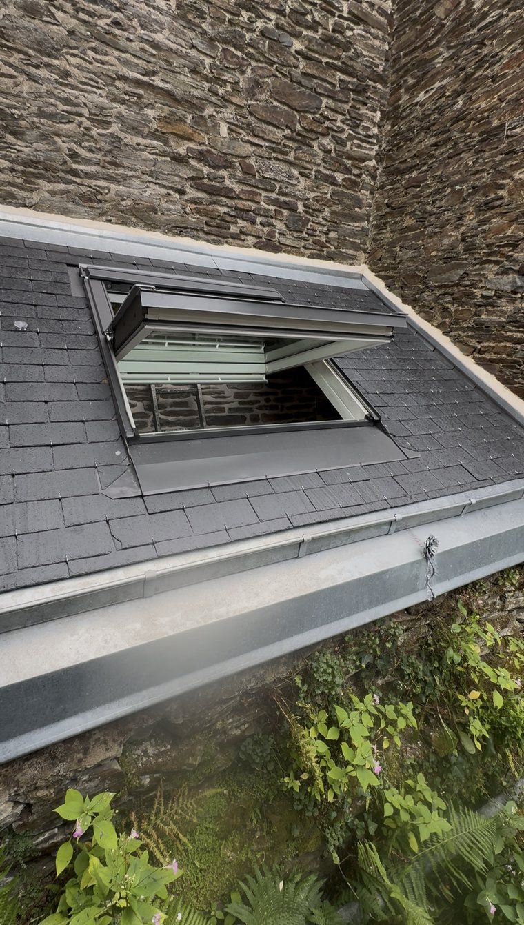 Fenêtre de toit VELUX ouverte sur une couverture en ardoise neuve, abergement en zinc et chéneau, entre deux murs de pierre à Saint-Malo