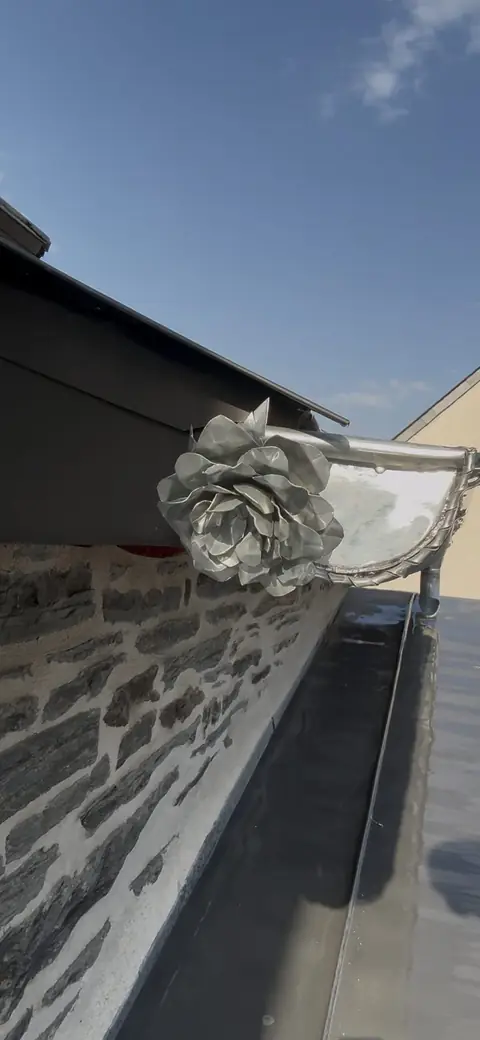Gros plan sur une rose en zinc aux pétales façonnés à la main, soudée sur la naissance d'une gouttière