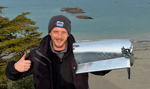 Willy Lambert, fondateur des Couvreurs Malouins, tient le membron d'angle en zinc et sa rose soudée, devant la plage de Saint-Lunaire