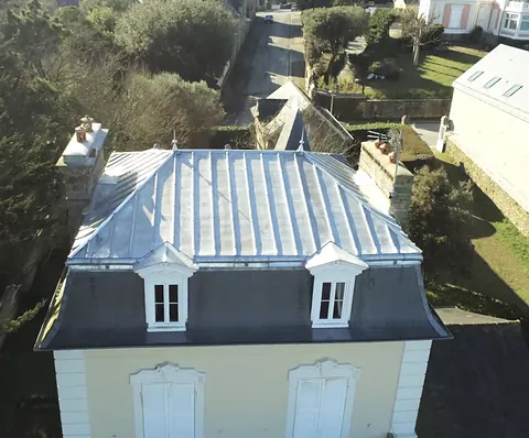 Vue au drone de la villa à la Mansart terminée, côté rue : terrasson en zinc naturel à tasseaux, épis aux angles, brisis en ardoise, deux lucarnes